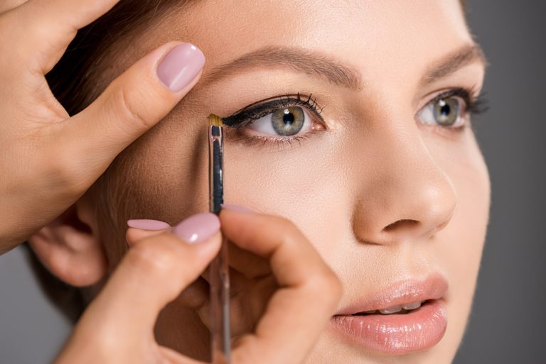 cropped shot of makeup artist applying eyeliner on models face on grey backdrop