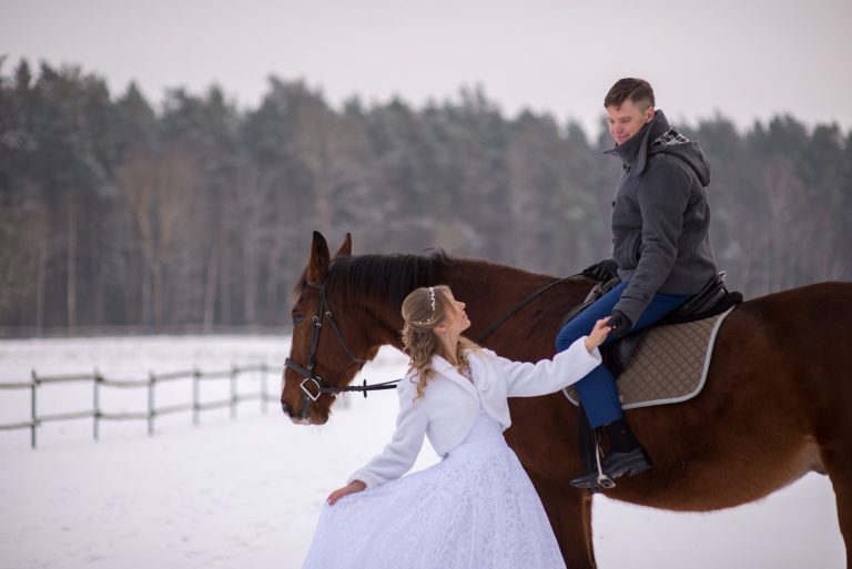 groom on brown horse and beautiful bride in a white dress standing on a snowy field, winter wedding