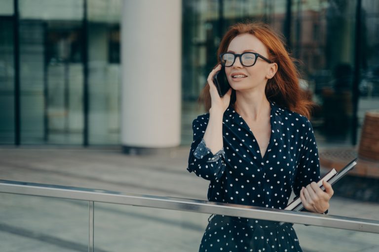 Thoughtful redhead woman wears spectacles polka dot dress has telephone conversation